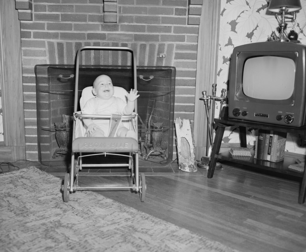 Old black and white image of baby in stroller in front of fireplace by television